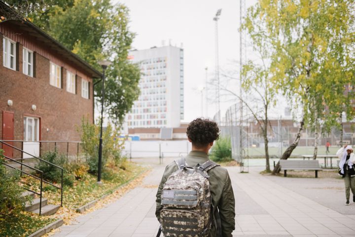 Ett barn med ryggsäck går längs en gångväg mellan ett tegelhus och höstlöviga träd, med ett högt vitt hus synligt i bakgrunden.
