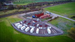 Flygfoto över batteripark och transformatorstation omgiven av gröna fält och järnväg. Foto.