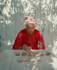 Nina Korhonen, Patience, Anna Amerikan mummu, 1993-99  - Äldre kvinna med röd knytblus och papiljotter lägger patience vid bord.