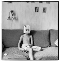 Svartvit bild av pojke som sitter med bar överkropp i soffa och har en pappåse med utklippta ögon på huvudet.