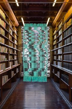 En väv med abstrakt mönster i turkos och grå, hänger mellan bokhyllor i ett bibliotek.