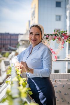 Kvinna står på en balkong i ett lägenhetshus och kollar in i kameran