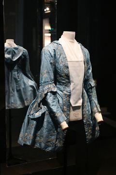 Från Textilmuseets samling: Sidenjacka med silverbroderier, 1770-1780-tal. Från Torpa stenhus, Länghem sn, Kind hd. I Textilmuseets samling finns inget större plagg bevarat av broscherat siden. Däremot finns några sidenplagg som i utställningen visar på modet under 1700-talet och som likväl hade kunnat varit sydda av broscherat siden.  Just det här är en så kallad Brunswick, en jacka med huva och lösärmar. Den här typen av jacka hade antingen en separat väst eller en bröstlapp fram med knäppning som idag tyvärr inte finns bevarad till detta exemplar. Till jackan bars en matchande kjol och dräkten användes som resdräkt.