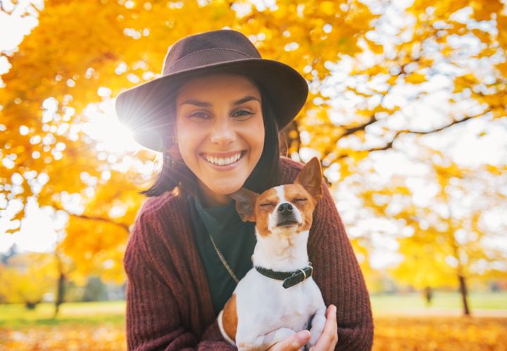 En kvinna i svart hatt med en liten vitbrun hund i famnen. I bakgrunden ett träd med gula höstlöv.