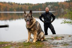 Årets polishund 2026 Sid sitter vid strandkanten till en sjö och i bakgrunden står hans förare Jesper Wretman.
