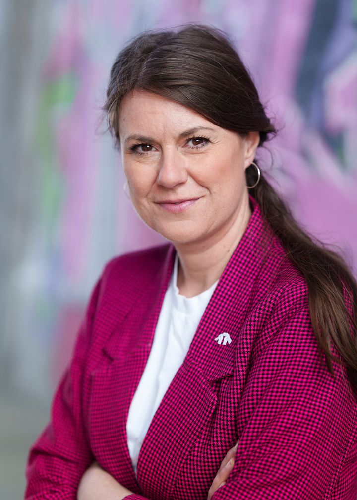 Kvinna med röd och svart-rutig kavaj och vit T-shirt tittar in i kameran och har armarna i kors