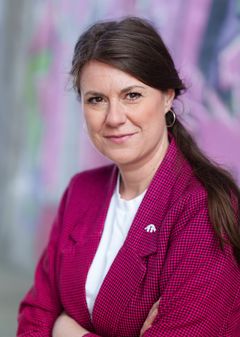 Kvinna med röd och svart-rutig kavaj och vit T-shirt tittar in i kameran och har armarna i kors