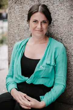 En person i svarta kläder och grön tunn skjorta sitter mot en stenvägg med armarna i knäet och tittar in i kameran