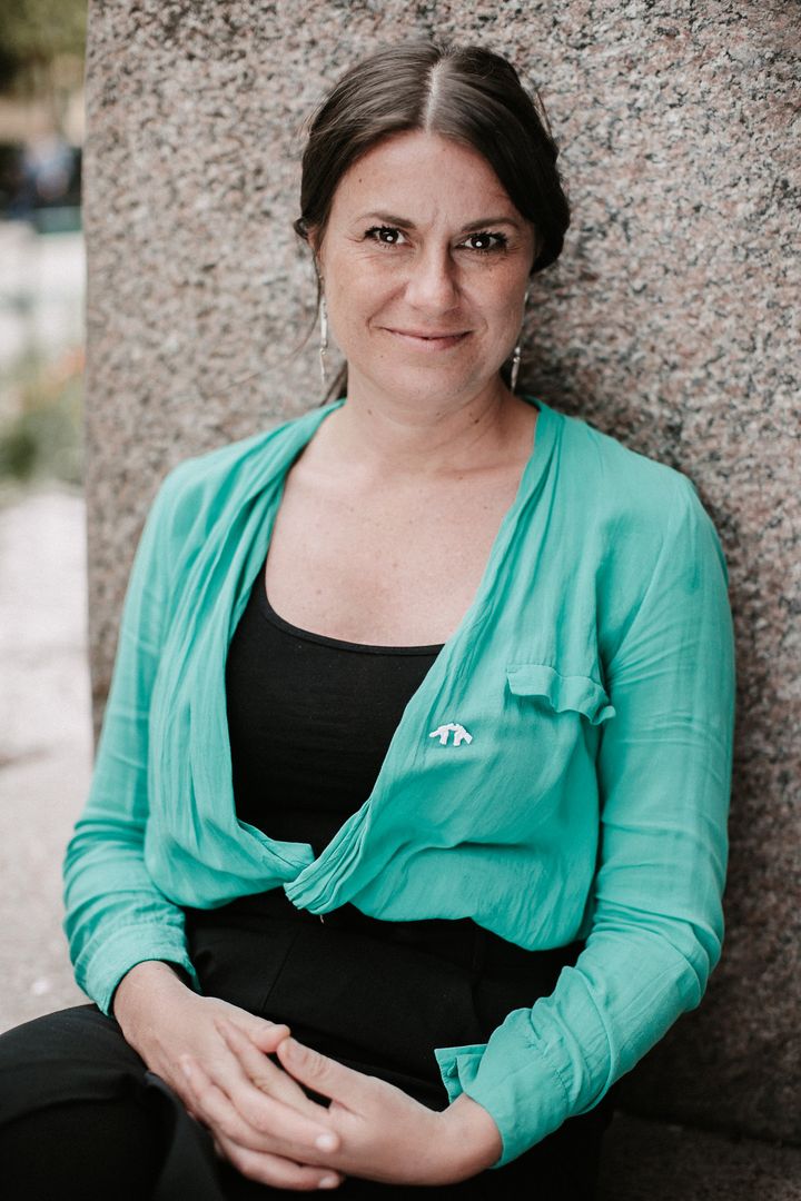 En person i svarta kläder och grön tunn skjorta sitter mot en stenvägg med armarna i knäet och tittar in i kameran