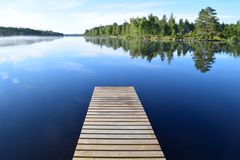 Brygga av trä som sträcker sig ut över en spegelblank sjö med skog i bakgrunden.
