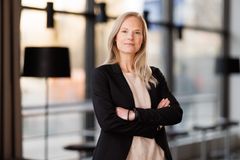 Bild på en kvinna med blont hår som står med armarna i kors, klädd i en svart kavaj, i en kontorsmiljö.