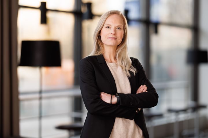 Bild på en kvinna med blont hår som står med armarna i kors, klädd i en svart kavaj, i en kontorsmiljö.