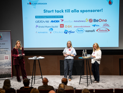 Tre personer på scenen under Blodcancerdagen med bild på de sponsrande företagens logotyper på väggen bakom