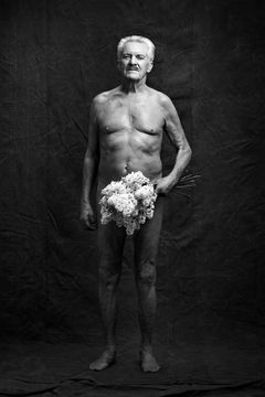 Äldre barbröstad man i mustasch som står upp och håller en blombukett i ena handen.