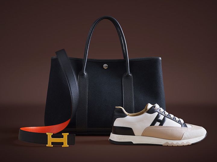 Svart handväska, vit och beige sneaker, och svart bälte med orange insida och guldfärgad H-spänne mot brun bakgrund.