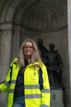 Bild på en kvinna i en gul reflexjacka stående framför en staty.