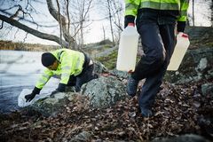 Två personer i gula reflexjackor fyller plastbehållare med vatten vid en sjö.