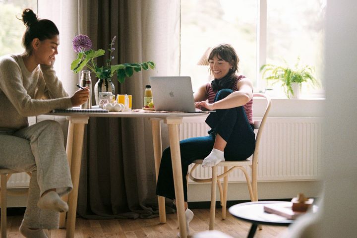 Två unga kvinnor sitter vid ett runt matbord och pluggar med laptop och papper/penna.