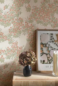 Tapet med små röda blommor och grönt bladverk på ljus botten. Framför väggen står en träbyrå med en svart vas, hortensia och en inramad tavla.