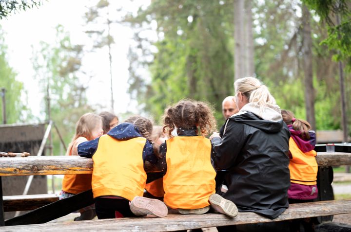 Barn och förskolepersonal sitter ner vid några bänkar utomhus i skogsmiljö
