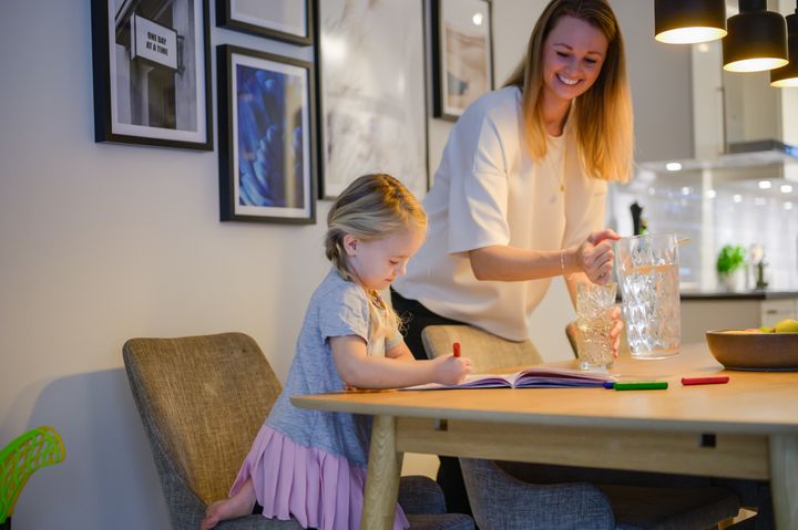 Ett barn ritar i en bok vid köksbordet. Bredvid står en vuxen med en vattenkanna och glas.